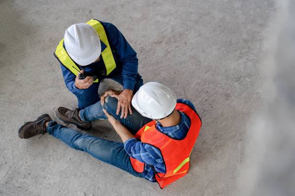A worker radios for help as his coworker sits on the ground with a knee injury