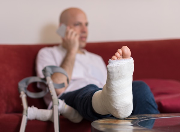 Young man with a broken ankle and a white cast on his leg, sitting on a red couch, on the phone with a workers' compensation attorney.