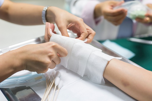 Close-up of a hand with a burn injury being wrapped with a white bandage by a nurse. The bandage is being carefully wrapped around the hand and secured in place, providing protection and support to the injured area. 