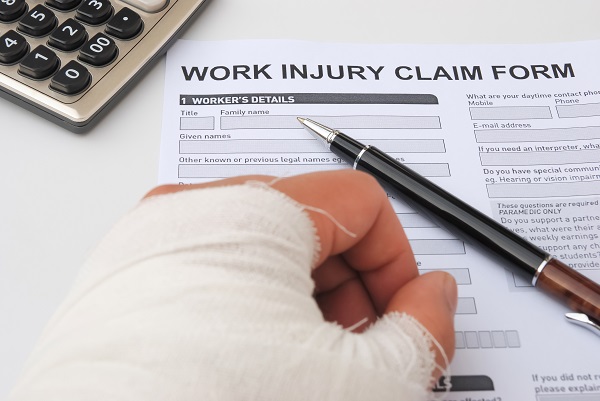 Person with a bandaged hand holding a pen while filling out a work injury claim form, with a calculator nearby on the desk.