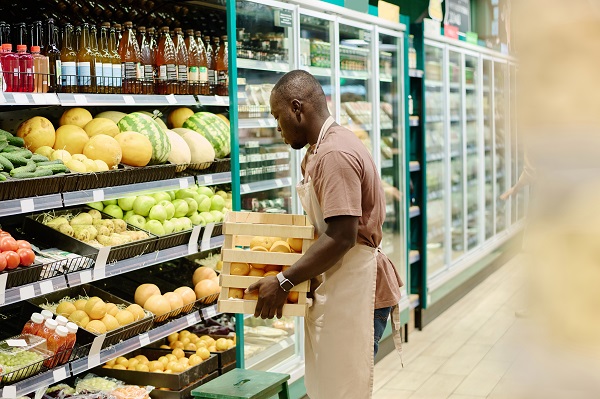 Grocery store worker in a beige apron lifting a wooden crate of lemons while stocking fresh produce on a shelf.