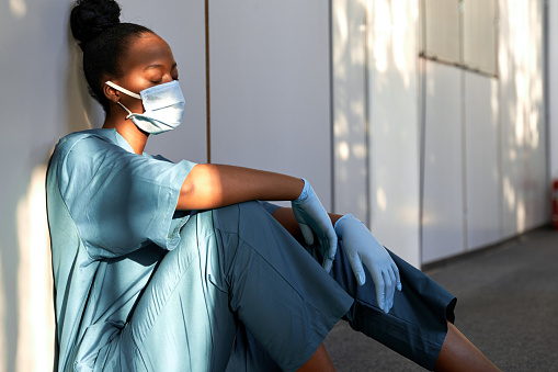 Exhausted female scrub nurse sits on hospital floor.