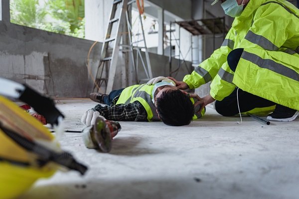 Construction worker lying on the ground after a fall, with a colleague learning over him for help, symbolizing a workplace injury incident on a job site.